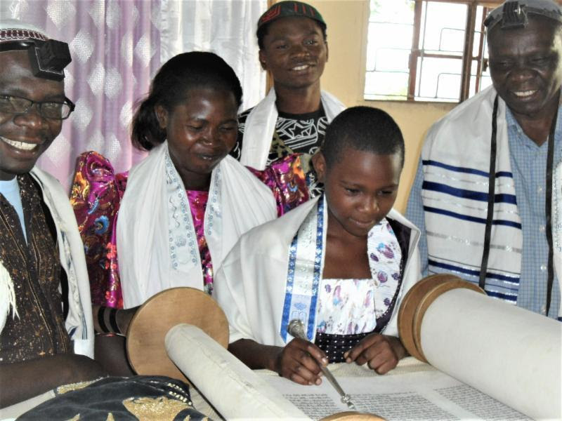 Ugandan child smiles and reads from a Torah scroll with family and community members watching.